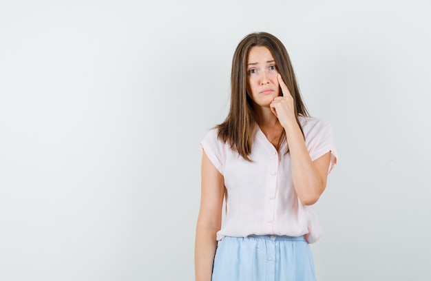 Young girl in t-shirt, skirt pointing finger at her eye and looking unhappy , front view.