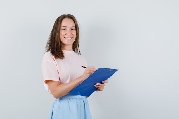 Young girl in t-shirt, skirt taking notes on clipboard and looking cheerful , front view.