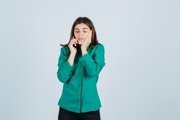 Young girl talking to phone, holding hand on cheek in green blouse, black pants and looking curious , front view.