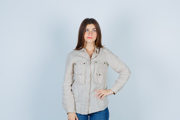 Free photo young girl thinking about something, with hand on hip in beige shirt, jeans and looking pensive , front view.