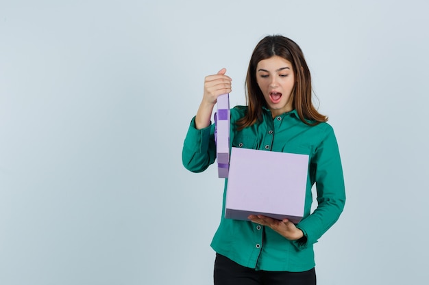 Young girl trying to open gift box in green blouse, black pants and looking surprised. front view.