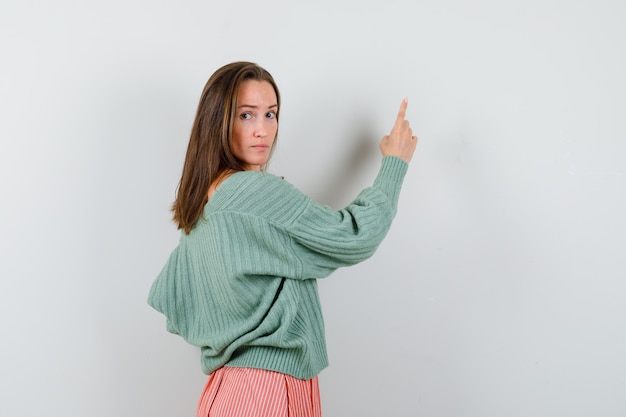 Free Photo young girl turning back, pointing at wall with index finger, looking at camera in knitwear, skirt and looking alluring , front view.