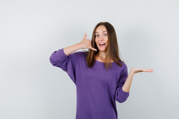 Young girl in violet shirt showing phone sign with palm spread aside and looking helpful , front view.