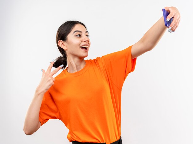 Young girl wearing orange t-shirt doing selfie using smartphone smiling showing v-sign standing over white wall