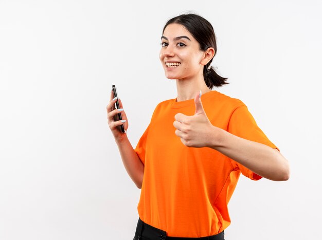 Young girl wearing orange t-shirt holding smartphone looking aside smiling cheerfully showing thumbs up standing over white wall