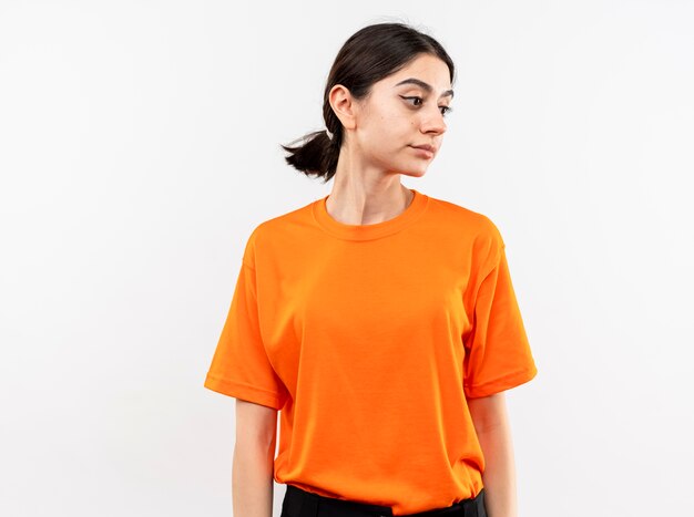 Young girl wearing orange t-shirt looking aside with sad expression standing over white wall