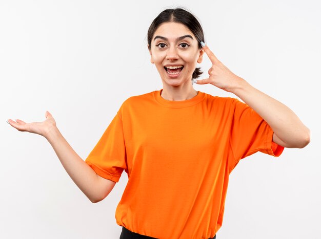 Young girl wearing orange t-shirt presenting something with arm showing rock symbol smiling cheerfully standing over white wall