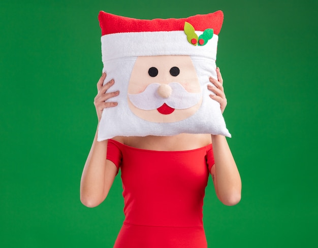 Free Photo young girl wearing santa hat holding santa claus pillow covering face with it isolated on green background