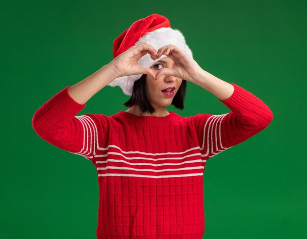 Young girl wearing santa hat looking at camera doing heart sign in front of eye isolated on green background