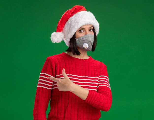 Free Photo young girl wearing santa hat and protective mask looking at camera pointing at side isolated on green background with copy space