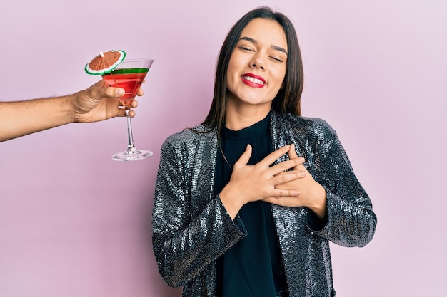 Free Photo young girl wearing sexy party jacket and hispanic arm offering cocktail smiling with hands on chest, eyes closed with grateful gesture on face. health concept.