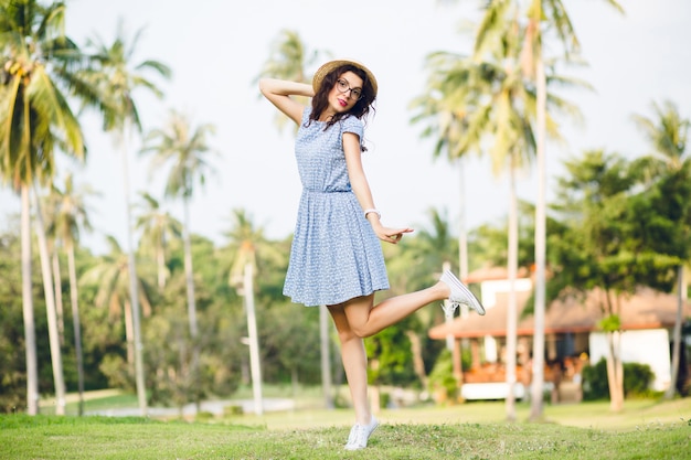 Young girl wearing sky-blue dress is standing on one leg on tip-toes in a park. Girl has straw hat and black glasses on.