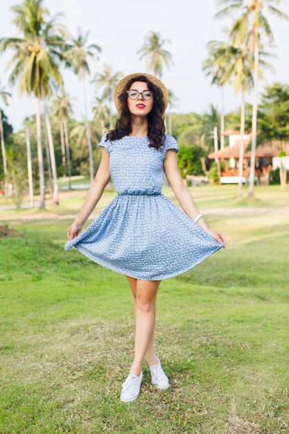 Young girl wearing sky-blue dress is standing on tip-toes in a park. Girl has straw hat and black glasses on