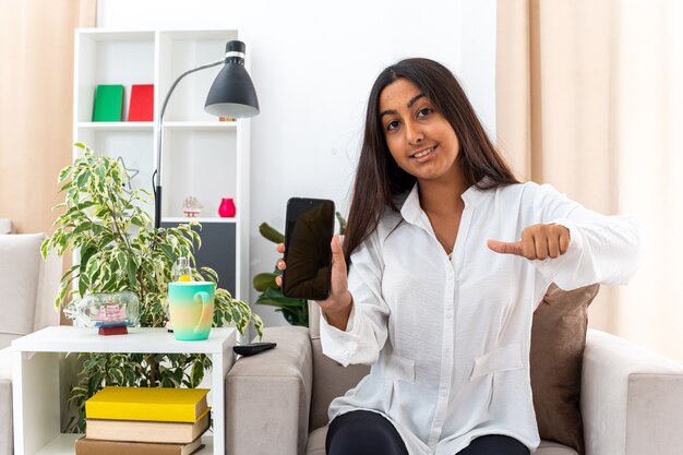 Young girl in white shirt and black pants holding smartphone showing thumbs up happy and cheerful sitting on the chair in light living room