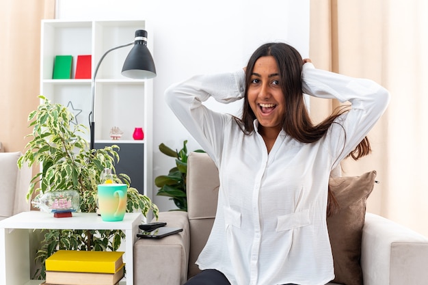 Young girl in white shirt and black pants looking at camera happy and excited holding hands on her head sitting on the chair in light living room