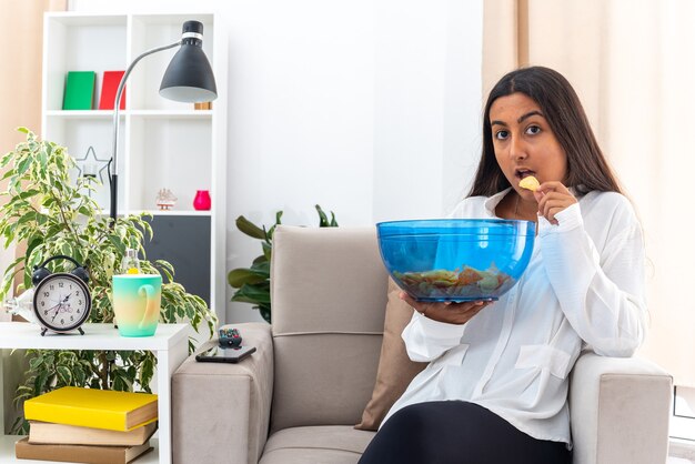 Young girl in white shirt and black pants with bowl of chips eating and sitting on the chair in light living room