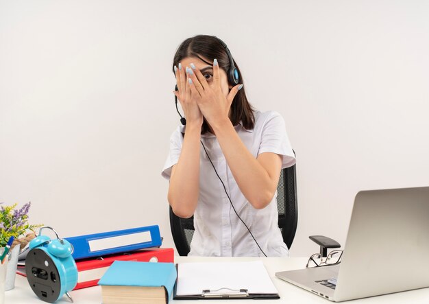 Young girl in white shirt and headphones, scared covering eyes with palms looking through fingers sitting at the table with folders and laptop over white wall