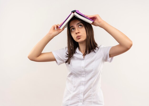 Young girl in white shirt holding open book over her head looking confused standing over white wall