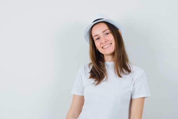 Young girl in white t-shirt, hat and looking cheerful , front view.