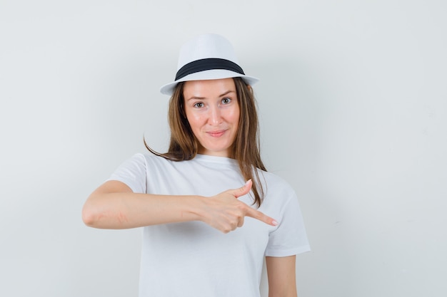 Free Photo young girl in white t-shirt, hat pointing down and looking confident , front view.