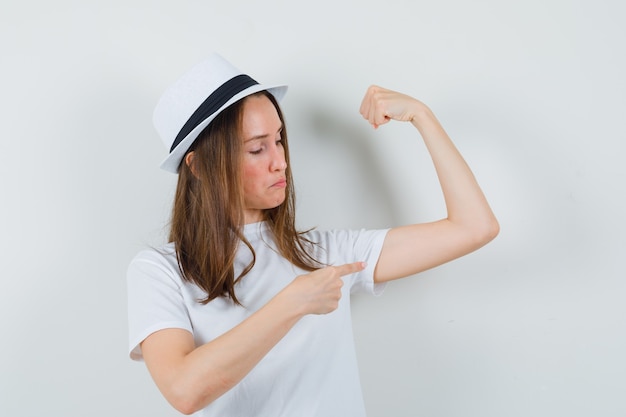 Free photo young girl in white t-shirt, hat pointing at muscles of arm and looking proud , front view.