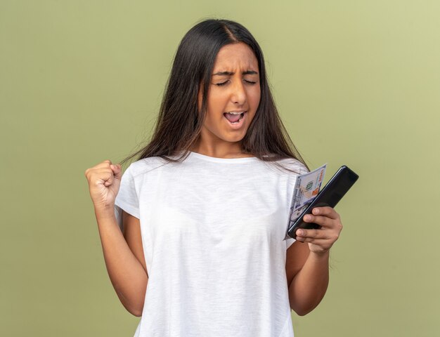 Young girl in white t-shirt holding smartphone happy and excited clenching fist standing over green