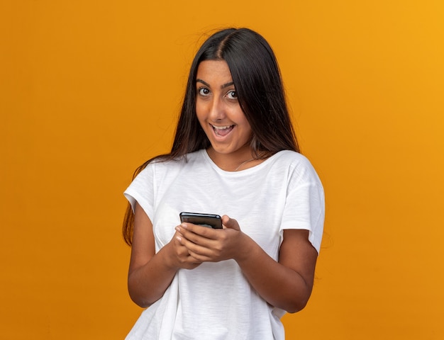 Young girl in white t-shirt holding smartphone looking at camera with happy face smiling cheerfully 