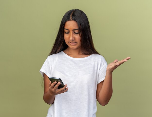 Young girl in white t-shirt holding smartphone looking at it being confused with arm raised 