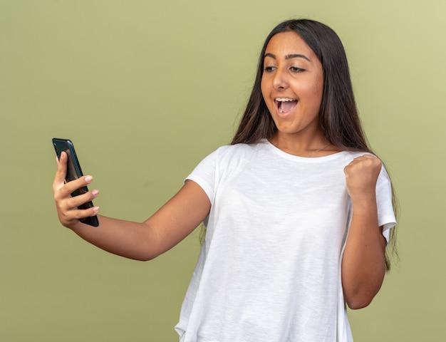 Young girl in white t-shirt holding smartphone looking at it happy and excited clenching fist standing over green