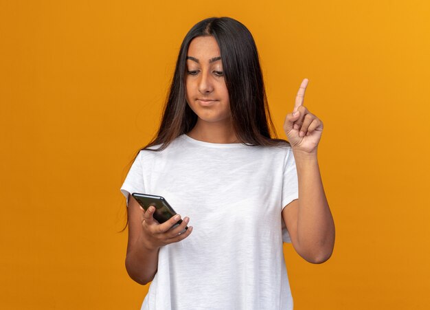 Young girl in white t-shirt holding smartphone looking at it with smile on smart face showing index finger having new idea standing over orange background