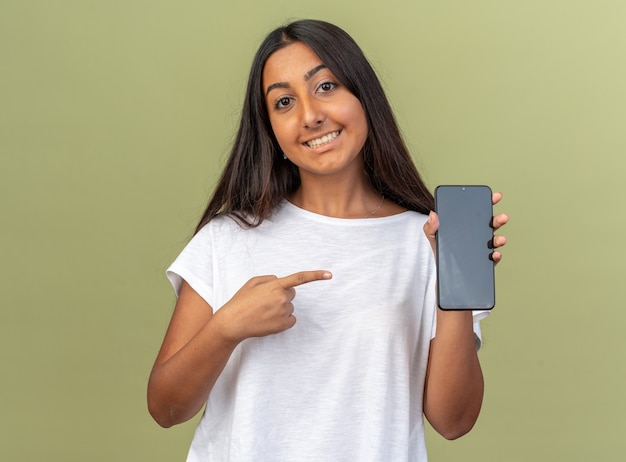 Young girl in white t-shirt holding smartphone pointing with index finger at it smiling cheerfully