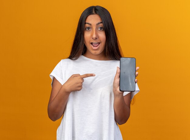 Young girl in white t-shirt holding smartphone pointing with index finger at it smiling