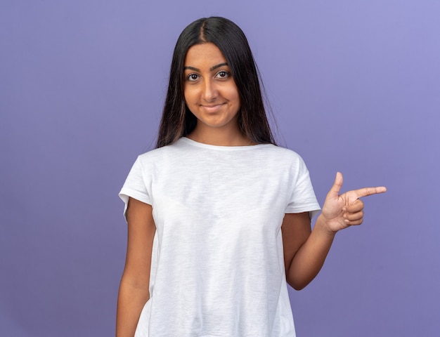 Young girl in white t-shirt looking at camera with smile on happy face pointing with index finger to the side standing over blue