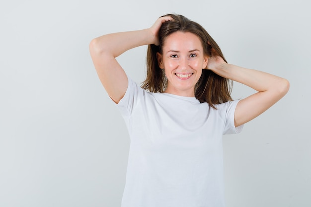 Young girl in white t-shirt posing with hands in hair and looking charming , front view.