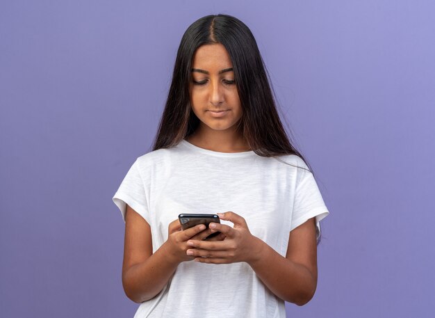 Young girl in white t-shirt writing text message using smartphone standing over blue