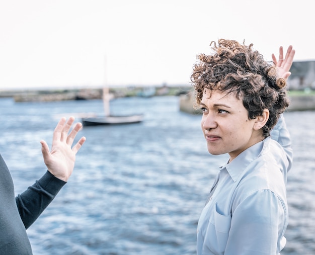 Young girl with curly hair and a piercing  nose arguing with her partner with expressive gestures with the  sea out of focus