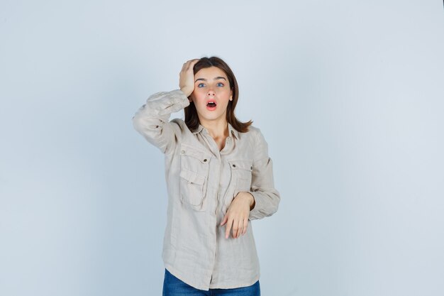 Young girl with hand on head, keeping mouth open in beige shirt, jeans and looking surprised , front view.