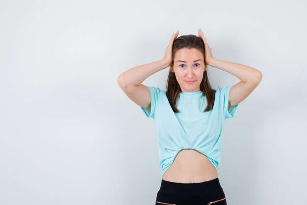 Young girl with hands on head in turquoise t-shirt, pants and looking wistful , front view.