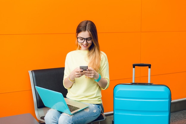 Free photo young girl with long hair and black glasses is sitting on chair on orange background. she wears jellow sweater with jeans. she has laptop on knees and suitcase near. she is typing on phone.