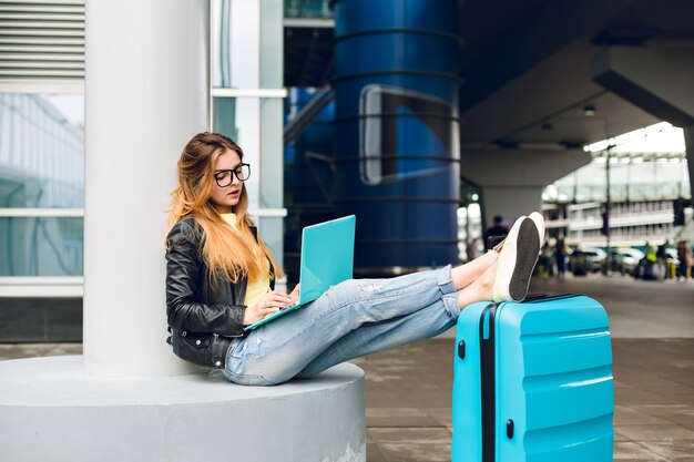 Young girl with long hair in black glasses is sitting outside in airport. She wears jeans, black jacket, jellow shoes. She put her legs on suitcase near. She is bored at typing on laptop.