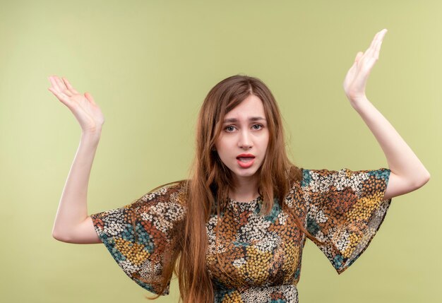 Free Photo young girl with long hair wearing colorful dress looking uncertain and confused raising hands, having no answer  