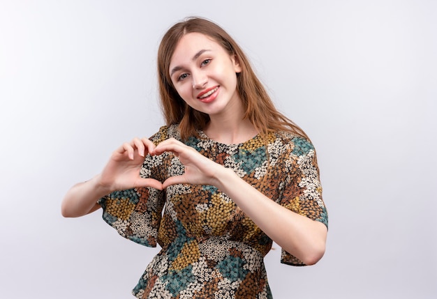 Free photo young girl with long hair wearing colorful dress positive and happy smiling broadly doing heart gesture with hand over chest