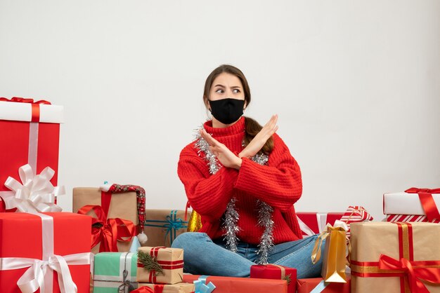 young girl with red sweater and black mask crossing hands sitting around presents on white