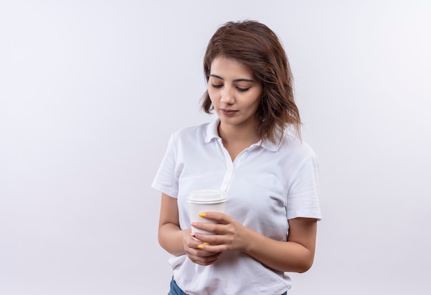 Young girl with short hair wearing white polo shirt holding coffee cup looking down with serious face 