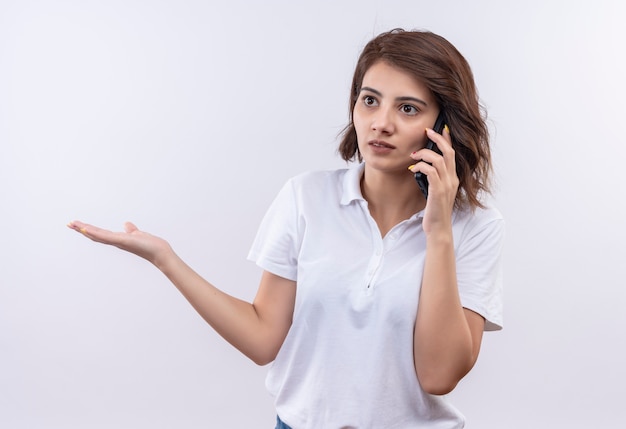 Young girl with short hair wearing white polo shirt looking confusedand very anxious while talking on mobile phone 