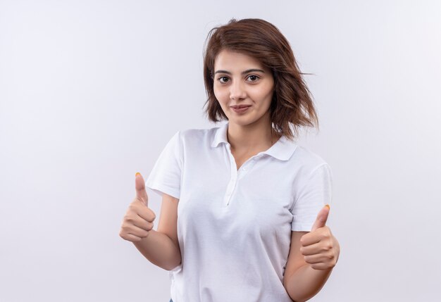 Young girl with short hair wearing white polo shirt smiling friendly showing thumbs up with both hands 