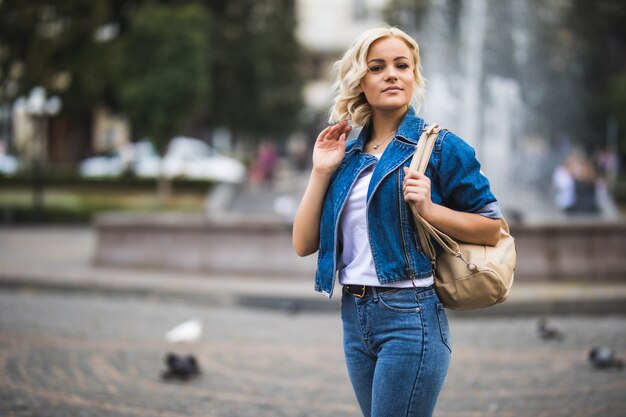 Young girl woman on streetwalk square fontain dressed up in blue jeans suite with bag on her shoulder in sunny day