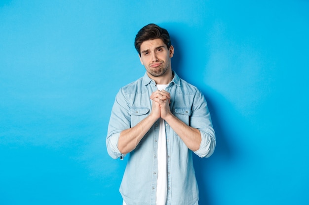 Free photo young guy begging for help, pouting and looking sad while asking for favour, need something, standing against blue background