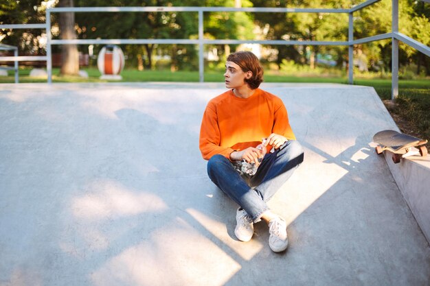 Young guy in orange pullover and jeans holding bottle of water in hand while thoughtfully looking aside at skatepark