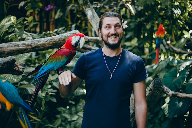 Free photo young guy posing in a zoo with a parrot in his hand, a bearded man and a bird